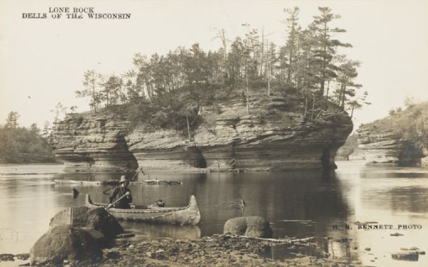 Photographic postcard view from shoreline of the rock formation in the Dells of the Wisconsin River. A man is paddling a canoe in the foreground. Printed on top of postcard: "Lone Rock, Dells of the Wisconsin."

