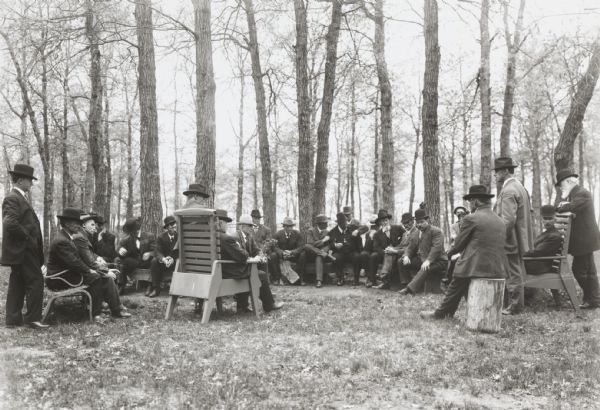 About two dozen men sit in chairs and on stumps in a clearing in a wooded area, having a meeting.