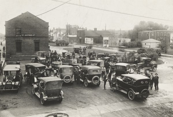 Elevated view of automobiles and trucks gathered in connection with the distribution of brook trout fish. The “milk” cans in sight presumably contain fingerling trout. The Antigo train station and platform is on the left behind the group of people gathered around the automobiles.
