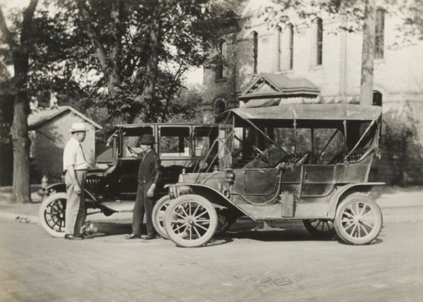 Two Ford automobiles, a 1910 touring car and a 1922 sedan, parked curbside. Two men stand in between the automobiles.
