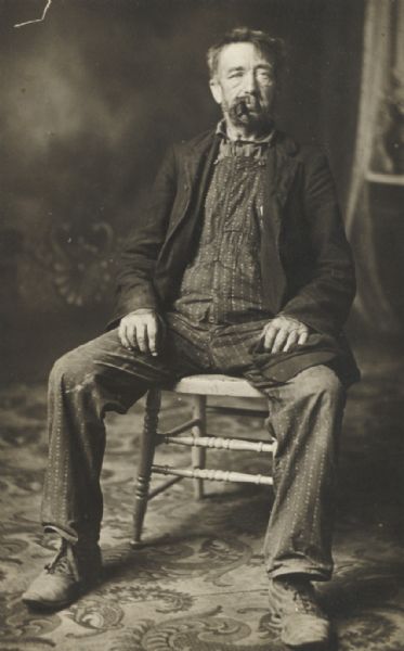 Studio portrait in front of a painted backdrop of a seated, unkempt man with a pipe in his mouth.