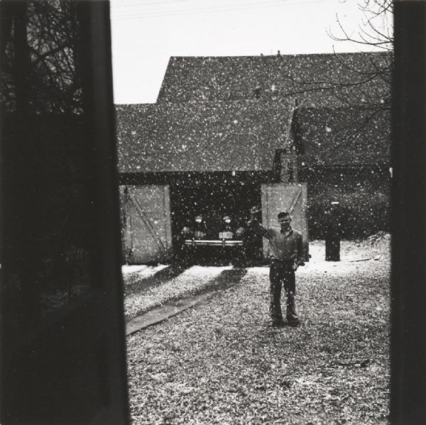 From a doorway framing the scene, a man is standing and waving in a yard during a snowstorm. Behind him an automobile is parked in a garage.