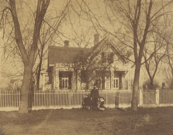View across road of the Clough house, residence of Mr. and Mrs. Willoughby. A man is standing and holding the hands of two young girls in front of the fence.