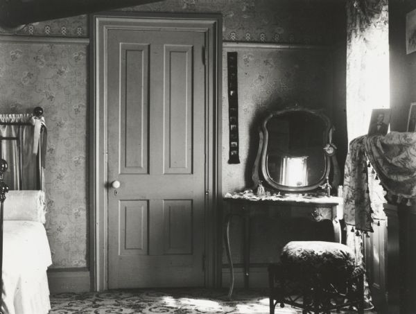 Interior of a bedroom, with part of a bed on the left, a closed door, and a dressing table with a mirror near a window and fireplace mantel. Otherwise unidentified.