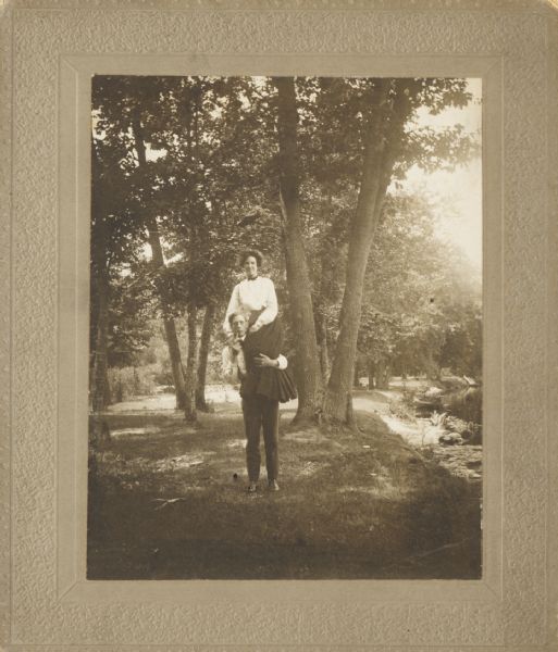 Young couple in a park or rural scene near a river. The man, “Mr. Fish” has lifted the young lady, Ida Langenberger, up onto his left shoulder. He is holding a pipe to his mouth with his right hand.
