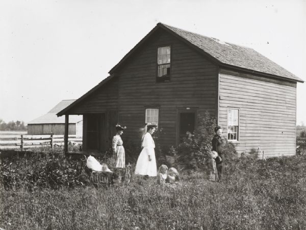 Three women and several children posed in the tall grass beside a small frame farmhouse.