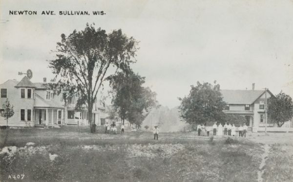 View across field of Newton Avenue. A group of people are posed in the field near trees, and houses are line the avenue on the left and right. Caption reads: "Newton Ave., Sullivan, Wis."