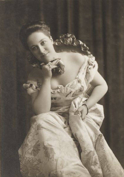 Unidentified lady, seated, wearing an off-the-shoulder short-sleeved ballgown, which appears to have fur decorating the collar and shoulders. She is resting her chin on her right hand on which she wears a number of rings.
