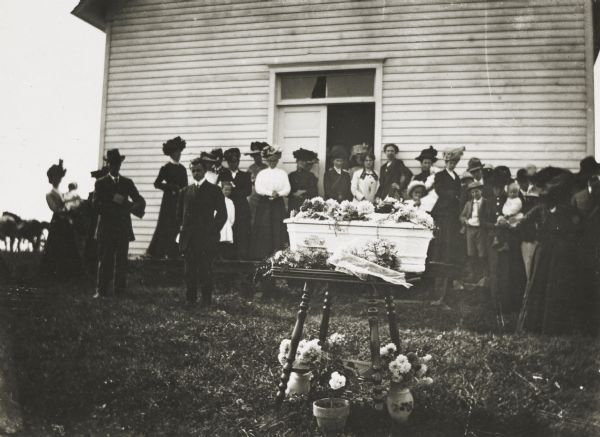 Group standing outdoors in the yard of a small clapboard church at the funeral of a small child. The child's white coffin, surrounded by mourners, stands on a small table in the foreground. There are flowers on top of the coffin, and potted flowers are on the ground underneath the table.

