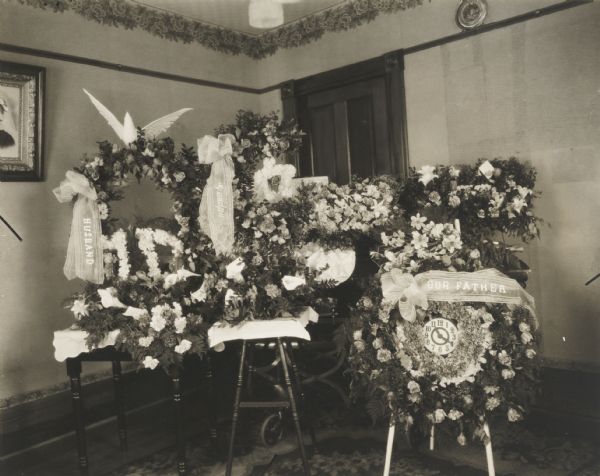 Floral decorations for a funeral, massed in the corner of a residential room. Printed on large ribbons adorning the decorations are the words: "Husband," "Our Father," and "Granpa." One wreath includes a clock in the center with the hands set at 11:20 a.m.
