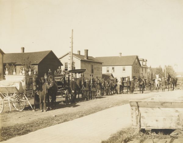 View towards vehicles in the street, including hearses, and horses from the Burton House Livery parked along the curb in front of storefronts. Men stand holding the bridles of some of the horses, and on the right in the back one man is posed sitting on a horse. 
