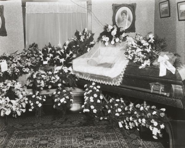 Deceased man's body lying in open veiled coffin, surrounded by flowers, in a residential interior.