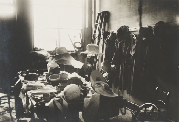 Apprentice milliner in training surrounded by hats.
