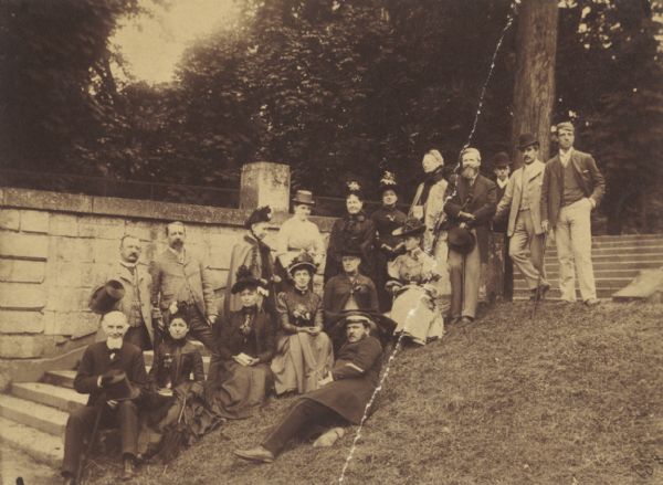Group of relatives and associates of John Johnston, posed sitting and standing on the steps of the estate. Some of the men wear bowler hats and hold canes, and two men are holding top hats. The woman wear hats and dresses, and two of them are wearing capes. A man is lying on the ground in front of the group wearing a cap.