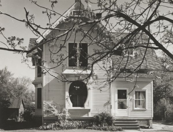 Frame residence with horseshoe arched front window. The tree in the foreground has blossoms on the branches.