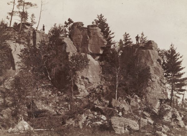 View looking up towards large rock formation with trees at Devil's Lake. A man is standing at the top of the rock formation on the right, and on the left a woman stands next to what is probably a dog.