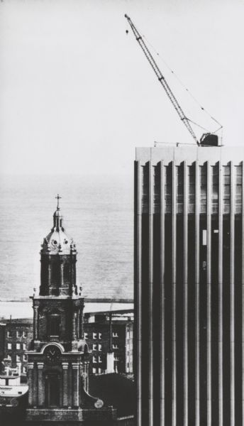 Elevated view of a detail on the Marshall & Ilsley Bank building, under construction. There is a crane on the roof. On the left is the tower of St. John's cathedral. In the background is Lake Michigan.