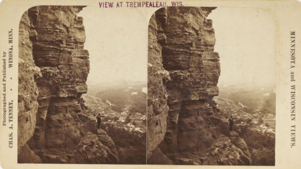 Stereograph view from the bluffs looking down over a town. A man is standing on the edge of the side of the formation, about halfway up, above treeline, and pointing to the right.