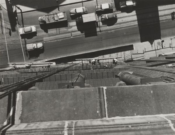 Overhead view looking down from a tall building at passing traffic and workmen on suspended scaffolding near the sidewalk.
