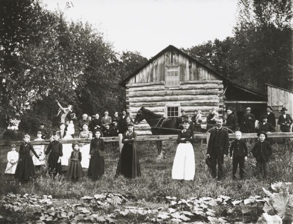 Large group, thirty or so, of men, women, children and infants posing standing in the yard of a house in front of and behind a wooden fence. In the center is a woman wearing a hat and glasses is sitting in a horse-drawn buggy. In the foreground is a garden with squash plants. Possibly a place near Irving, Wisconsin, between the Irving store, beyond the creek and Melrose.
