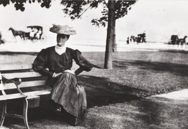 Mrs. Joseph Smith seated on a bench in Jackson Park. In the background are horse-drawn vehicles.