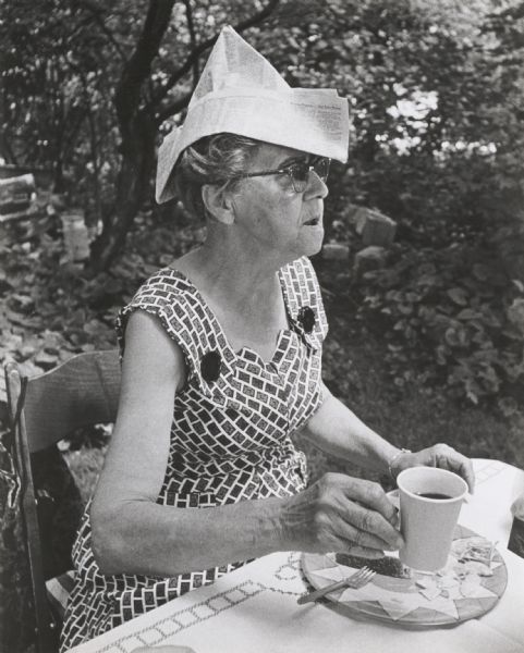 Middle-aged woman seated at an outdoor table with refreshments. She is wearing a hat made out of newspaper. The occasion is a card party arranged for older adults from Neighborhood House.