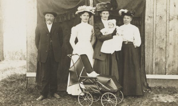 Unidentified posed group of an elderly man, and three women, with the one in the center holding an infant in her arms. The group is standing outdoors in front of a dark curtain with fringe that is hanging from the side of a wooden building. In front of the group is a baby carriage.