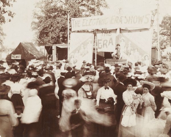 Crowd scene and circus barker in front of “The Electric Era Shows” tent, probably at a fair or carnival. A smaller tent in the background on the left has a sign for: "Palmist."