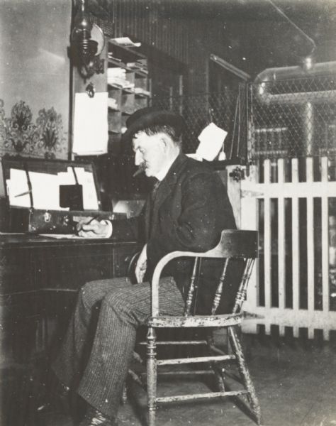 Man, seen from the side, seated working at a small desk or table and smoking a cigar. Behind him is a wooden gate and a metal fence.