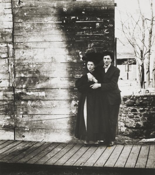 Young couple in moderately intimate pose against the end of a tall board fence on a boardwalk. A stone wall is next to the fence, and in the background on the right are trees and a building.