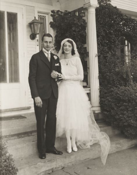 Wedding portrait of Mr. and Mrs. E. Randall Sears posed on front porch steps.  On June 19, 1928 Catherine McCaffrey married E. Randall Sears at the Christ Presbyterian Church, 2020 Chamberlain Avenue, Madison, Wisconsin.

According the June 19, 1929 issue of The Capital Times (p. 8), Miss McCaffrey wore "a formal gown of white satin, the bouffant skirt of which is longer in back than front, and a lace-trimmed veil falling from a closely fitted lace band headdress with orange blossoms on one side."  She carried "a shower bouquet of white roses and lilies of the valley."