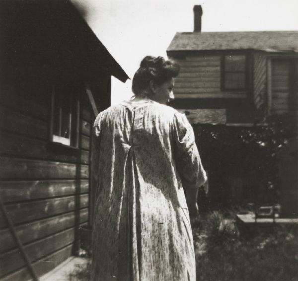 Woman wearing house dress, seen from behind, probably in the backyard of a rural residence.
