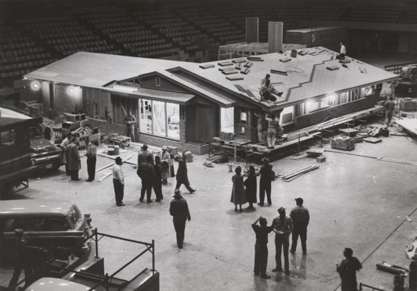Elevated view of demonstration “ranch” type frame residence under construction within an auditorium, presumably in Milwaukee, for some type of trade show. Construction workers are working on the roof. Men and women and parked vehicles are in the foreground.
