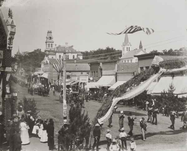 Elevated view of patriotic parade on Main Street, probably a Memorial Day Parade. Over the street, reaching from one sidewalk to the other, is an arch decorated with evergreen boughs, bunting and flags. Evergreen trees are also displayed along the sidewalks.