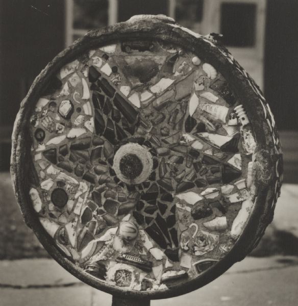 Homemade decorative star wheel set curbside. The object consists of an assortment of pottery sherds and other discarded bits set in cement within a circular rim.
