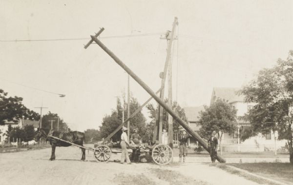 Photographic postcard of telephone company workmen erecting telephone pole with a horse-drawn derrick.