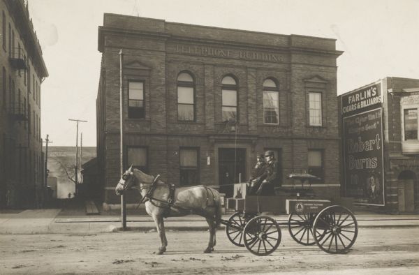 Wisconsin Telephone Company horse-drawn service and installation wagon in front of the Telephone Building. Two men sit in the wagon. A man stands in the doorway behind them. Above the doorway is a decorative lintel with a lion's head above a bell.
