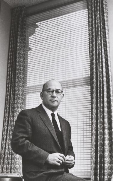 Executive portrait: man sitting on edge of desk in front of tall office window with curtains framing blinds.

