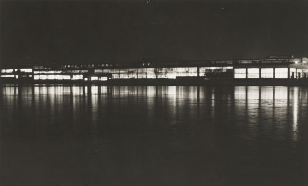 Long, low industrial building (factory?), probably in Green Bay, Wis., at night, from across the river, with reflections of interior lighting.