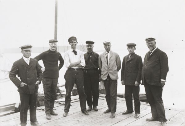 Seven men, standing informally, some wearing “yachting caps”, on a dock, presumably in Sturgeon Bay, Wis.