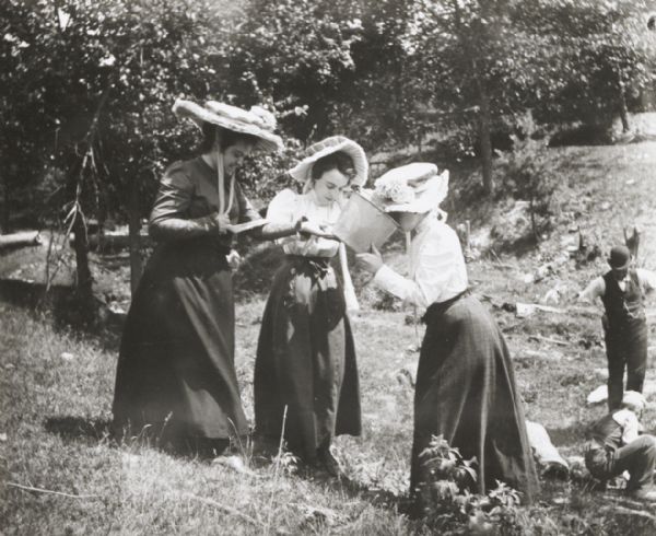 Three young women, perhaps at a picnic, with the woman on the right drinking from a bucket. Two men are behind them on the right.