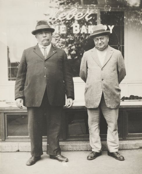 Two middle-aged men standing in front of the window of the Weber [Ho]me Bakery.