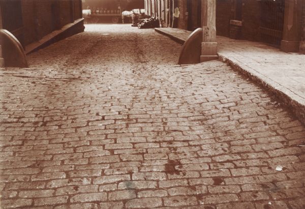 Paved driveway, cobblestones, leading to the loading platform of the Pabst Brewing Co. In the background, three horses stand near a wagon loaded with barrels. Brick buildings are on the left and right.
