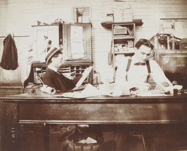 Two men in a small office, probably a railroad station. On the right is a map on the wall, and below it is a Herring Hall Marvin safe.