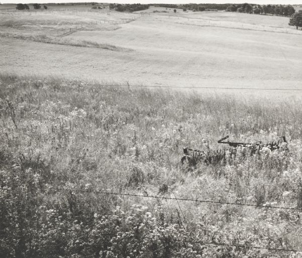View down slope across open fields over a barbed wire fence towards the horizon. The field is uncut in the foreground, obscuring an abandoned farm implement.
