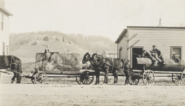 Men on wagons carrying large boiler, etc., posing in sham-battle horseplay, probably in Cross Plains. In the background is a bluff.
