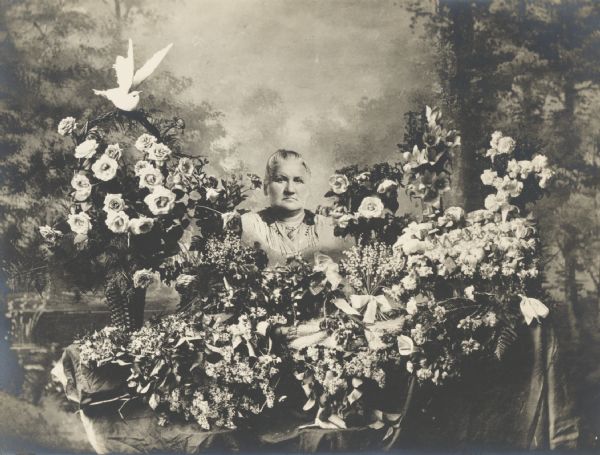 Memorial photograph, showing a draped coffin surrounded by floral offerings, among which appears a life-size head-and-shoulders photographic portrait of the deceased, an elderly woman.