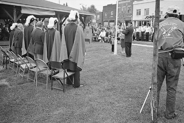 The flag raising during the Fourth of July ceremony, with a group of the local Knights of Columbus on the left. In the foreground on the right is a photographer with a tripod.