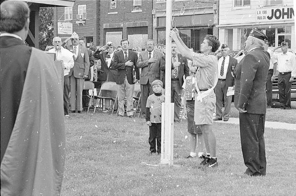 As a boy scout raises the flag, the people in attendance at the Park Falls Fourth of July celebration recite the Pledge of Allegiance. Congressman David R. Obey (in the dark jacket) stands to the left of the flag pole.