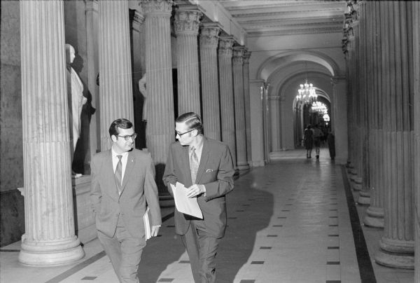Congressman David R. Obey with his administrative assistant Lyle Stitt walking down a hall in the United States Capitol building. Statues are along the wall between the stone columns.